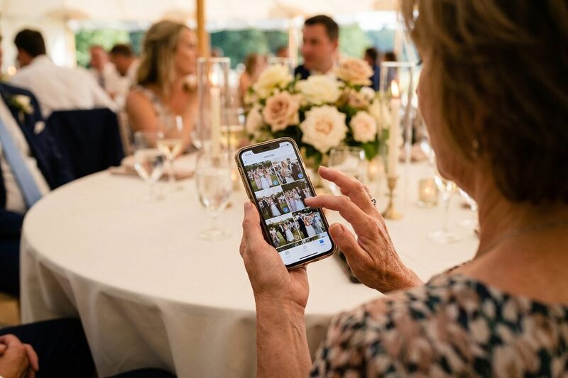 Over the shoulder of a wedding guest selecting a few photos from her camera roll to share at a reception table