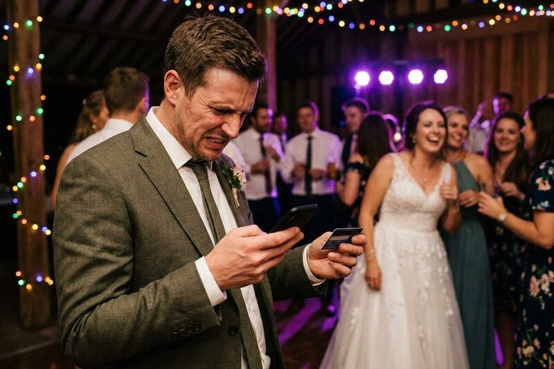 A groom grimacing at his phone with a bank card in his hand at the edge of the dance floor while the bride laughs behind him