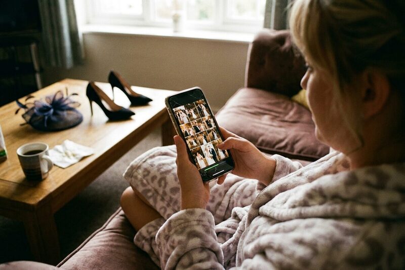 A wedding guest in a dressing gown uploading wedding photos on her phone the morning after, with her heels and fascinator on the coffee table