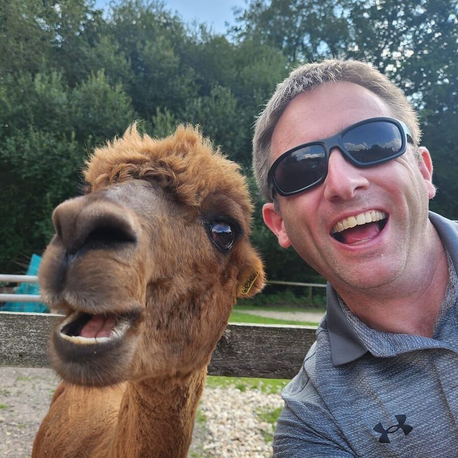 Alex Buckland in sunglasses taking a laughing selfie with an alpaca