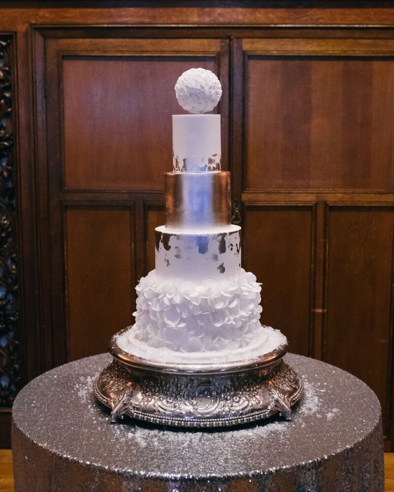 A four-tier white wedding cake with silver leaf, a sugar ruffle bottom tier and a sugar ball on top, on a silver stand in a wood-panelled room
