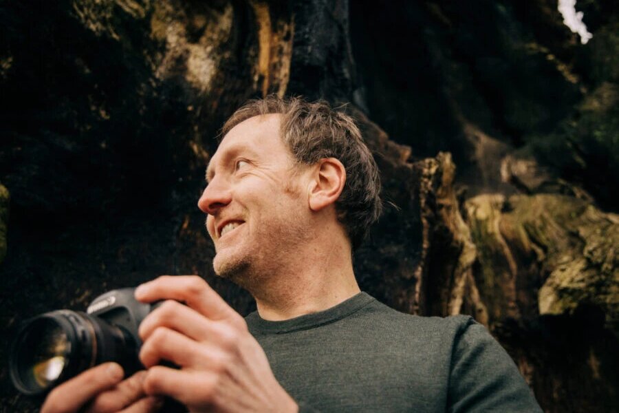 Photographer Darren Cresswell smiling and holding a camera in front of a mossy old tree