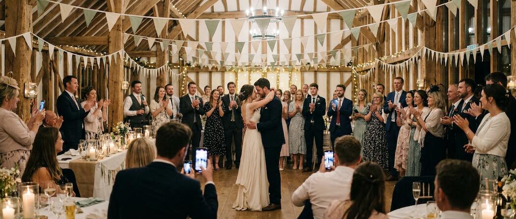 Wedding guests standing around the newlyweds during their first dance, several holding up phones to photograph it