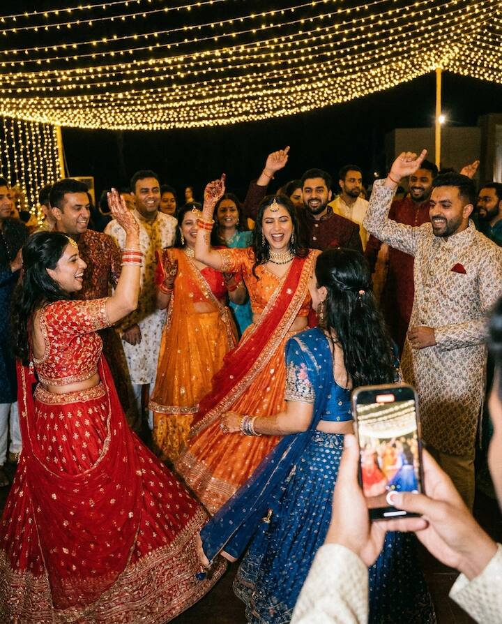 Guests dancing in colourful lehengas at a sangeet under string lights while a friend films on a phone